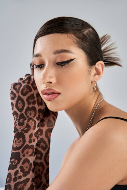 portrait de belle femme asiatique rêveuse en gants imprimés animaux et accessoires argentés, coiffure tendance et maquillage audacieux sur fond gris, printemps élégant, tournage de mode - Photo, image