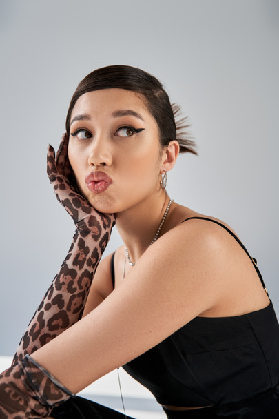 portrait of fashionable asian woman with bold makeup and brunette hair pouting lips and looking away on grey background, spring fashion, black dress, animal print gloves, silver accessories - Foto, immagini