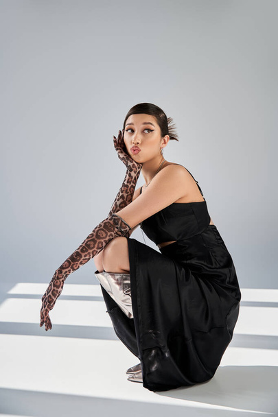 attractive asian woman in black elegant dress, animal print gloves and silver boots sitting on haunches, blowing kiss and looking away on grey background with lighting, spring fashion photography - Photo, Image