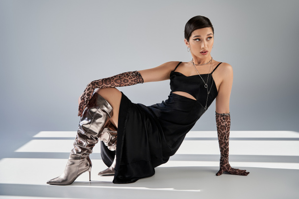 full length of trendy asian woman with bold makeup, in silver boots, black strap dress and animal print gloves posing and looking away on grey background with lighting and shadows, spring fashion - Photo, Image