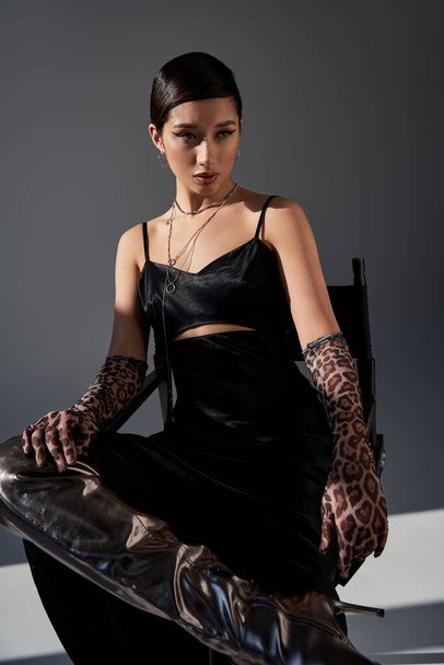 spring fashion concept, young asian woman with bold makeup, brunette hair, black strap dress, animal print gloves and silver boot sitting on chair on grey background with lighting, generation z - Foto, Bild