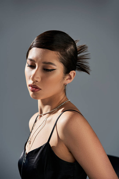 portrait of appealing asian woman with brunette hair, bold makeup, trendy hairstyle, in black strap dress and silver accessories posing in lighting on grey background, spring fashion concept - Φωτογραφία, εικόνα