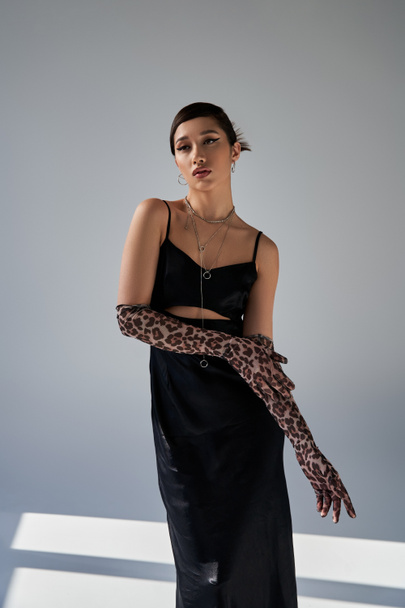 graceful asian woman with bold makeup and brunette hair standing in black strap dress and animal print gloves while looking away on grey background with lighting, stylish spring, fashion shoot - 写真・画像