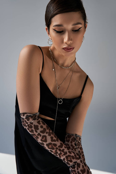 fashionable asian woman with bold makeup, brunette hair, silver necklaces and animal print gloves posing in black strap dress on grey background, stylish spring, generation z - Photo, Image