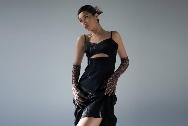 fashionable spring, mesmerizing asian woman in black and elegant strap dress, animal print gloves, with silver accessories and bold makeup looking away on grey background, generation z - Φωτογραφία, εικόνα