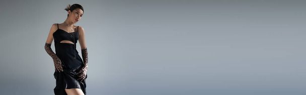 jugendliche und sinnliche asiatische Frau mit brünetten Haaren posiert in schwarzem Trägerkleid und Animal-Print-Handschuhen vor grauem Hintergrund, trendiges Frühjahrskonzept, Generation Z, Banner - Foto, Bild