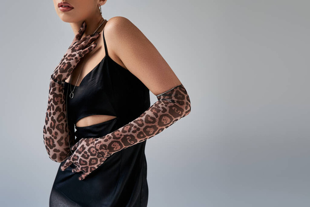 partial view of fashionable woman in black and elegant strap dress, silver necklaces and animal print gloves touching neck on grey background, trendy spring, generation z - Foto, afbeelding