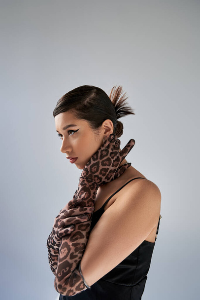 graceful asian woman with trendy hairstyle and bold makeup posing in black strap dress and animal print gloves while looking away on grey background, spring fashion photography - Fotó, kép