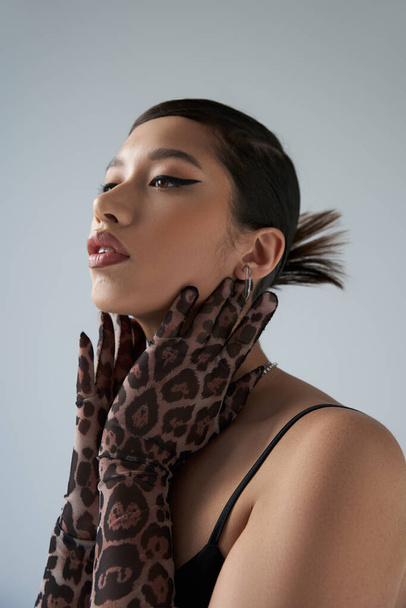 youthful style, spring fashion, young asian woman with brunette hair and bold makeup, wearing black dress and animal print gloves, touching neck, expressive, on grey background  - Foto, immagini
