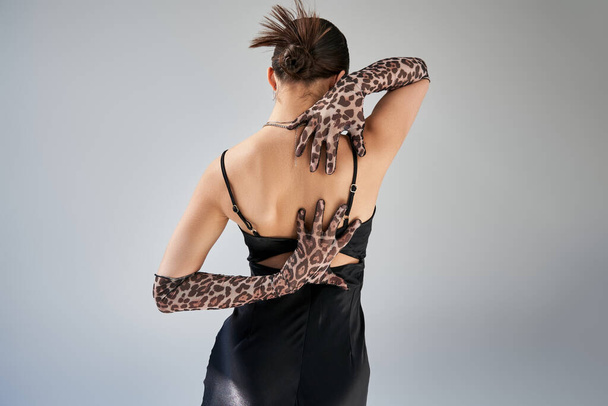 back view of young brunette woman with trendy hairstyle posing with hands behind back on grey background, black strap dress, animal print gloves, trendy spring, fashion photography - Valokuva, kuva
