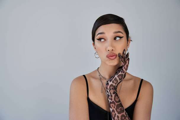 portrait of fashionable asian woman with skeptical face expression looking away on grey background, brunette hair, silver accessories, animal print glove, black dress, trendy spring concept - Photo, Image