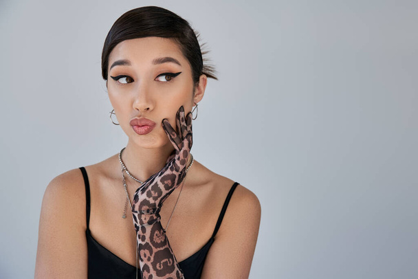 portrait of young asian woman in black strap dress, silver necklaces and animal print glove, with thoughtful and skeptical face expression looking away on grey background, spring fashion photography - Photo, Image