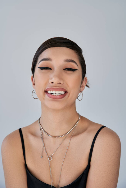 Porträt einer jungen und fröhlichen asiatischen Frau mit brünetten Haaren, fettem Make-up, silbernen Halsketten und Ohrringen, die auf grauem Hintergrund in die Kamera blickt, Konzept der Generation Z, Frühjahrsstil - Foto, Bild