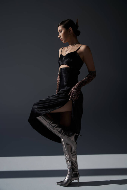 full length of graceful asian woman in black elegant dress, silver boots and animal print gloves standing on dark grey background with lighting, stylish spring, fashion shoot - Φωτογραφία, εικόνα