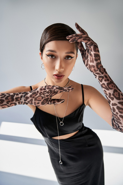 trendy spring, youthful style, brunette asian woman in black strap dress, animal print gloves and silver accessories holding hands near face and looking at camera on grey background with lighting - Foto, Bild