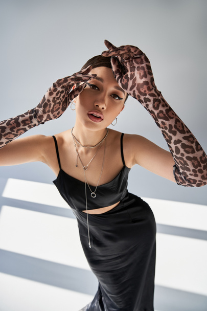 high angle view of asian woman in spring outfit, with expressive gaze looking at camera on grey background with lighting, silver accessories, black strap dress, animal print gloves, fashion shoot - Φωτογραφία, εικόνα