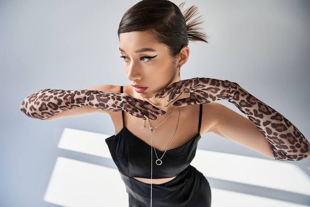 high angle view of elegant asian woman with bold makeup, wearing black strap dress and animal print gloves, holding hands under chin on grey background with lighting, spring fashion concept - Photo, Image