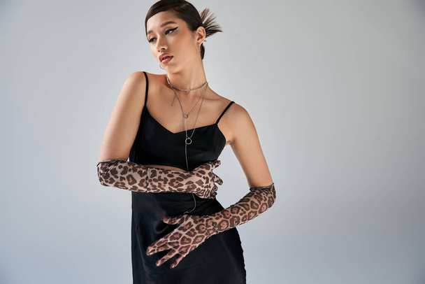 attracive asian woman with bold makeup and brunette hair posing in silver necklaces, black strap dress and animal print gloves on grey background, high fashion, spring style - Photo, Image