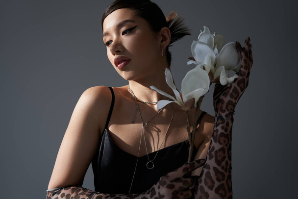 encantadora mujer asiática con maquillaje audaz y cabello moreno sosteniendo flor de orquídea blanca mientras posa en vestido de correa negro sobre fondo gris oscuro, fotografía de moda de primavera - Foto, imagen