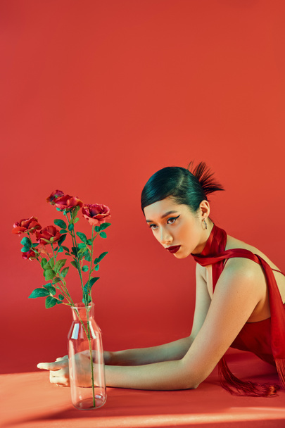 young and elegant asian woman in stylish neckerchief and dress, with bold makeup and brunette hair laying near glass vase with roses on red background with lighting, spring fashion, generation z - Photo, Image