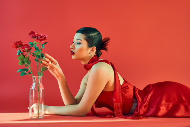 zijaanzicht van charmante aziatische vrouw met brunette haar en gedurfde make-up, in elegante jurk en halsdoek liggen en aanraken rozen in glazen vaas op rode achtergrond met verlichting, lente mode concept - Foto, afbeelding