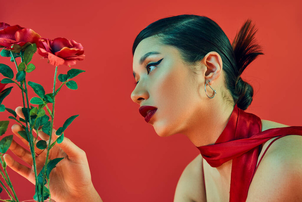 side view of mesmerizing asian woman with brunette hair, bold makeup and trendy hairstyle touching green leaves of roses on red background, generation z, spring fashion concept - Photo, Image