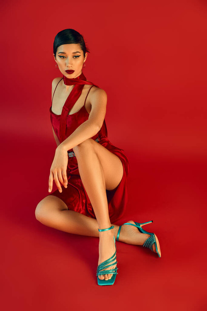 full length of elegant asian woman in stylish spring outfit sitting in stylish pose on red background, brunette hair, strap dress, neckerchief, turquoise sandals, generation z fashion - Photo, Image