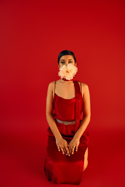 spring fashion concept, young asian woman with brunette hair and bold makeup, wearing elegant dress and neckerchief, holding white peony in mouth while kneeling on red background - Photo, Image