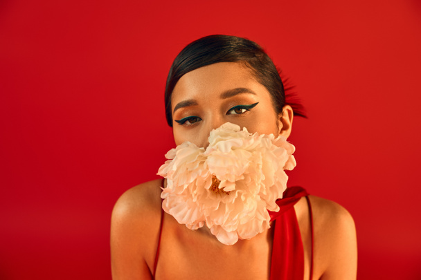portrait of youthful and sensual asian fashion model with bold makeup and brunette hair holding white peony in mouth and looking at camera on vibrant red background, gen z fashion, spring style - Foto, Imagem