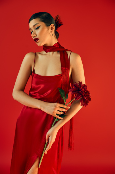 youthful and sensual asian woman with bold makeup and brunette hair, in glamour dress and neckerchief posing with burgundy peony on red background, trendy spring, gen z fashion - Foto, imagen