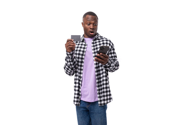 překvapený atraktivní 30 letý africký chlap oblečený v kostkované černé a bílé košile používá internetové bankovnictví na smartphone na pozadí s kopírovacím prostorem. - Fotografie, Obrázek