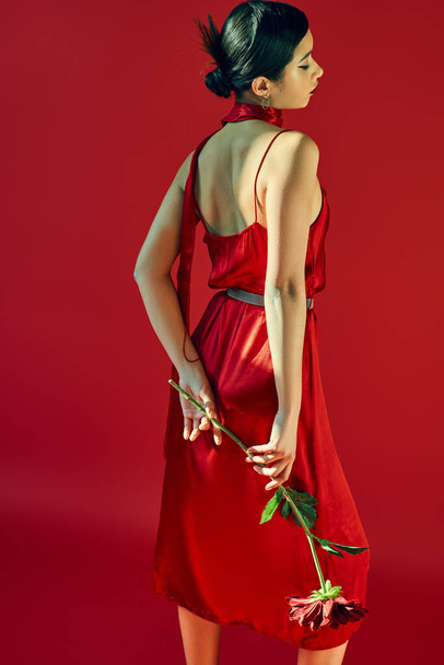 jovem e elegante asiático mulher com morena cabelo e moderno penteado de pé em elegante pose com borgonha peônia no vermelho fundo, na moda primavera, geração z - Foto, Imagem