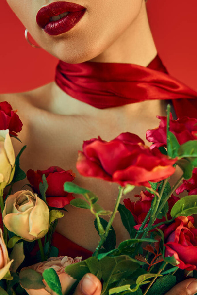 κομψό έννοια άνοιξη, περικοπή άποψη του νεαρού μοντέλου μόδας με φωτεινά χείλη και μαντήλι ποζάρουν με μπουκέτο από φρέσκα τριαντάφυλλα σε κόκκινο φόντο, φωτογράφηση μόδας, γενιά z - Φωτογραφία, εικόνα