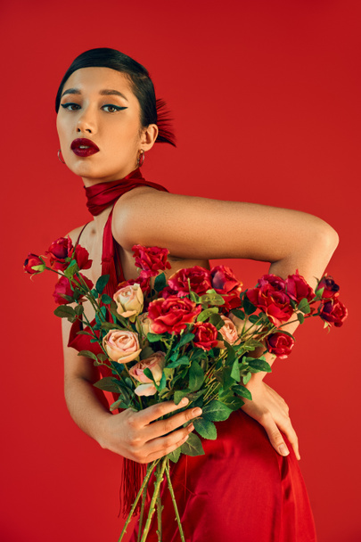 young and seductive asian woman with bold makeup, in trendy dress posing with hand on hip and roses while looking at camera on red background, trendy spring, fashion shoot - Φωτογραφία, εικόνα