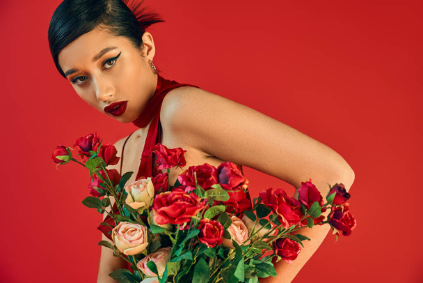 youthful and mesmerizing asian woman with bold makeup, brunette hair and seductive gaze looking at camera near bouquet of fresh roses on red background, trendy spring, generation z - Photo, Image