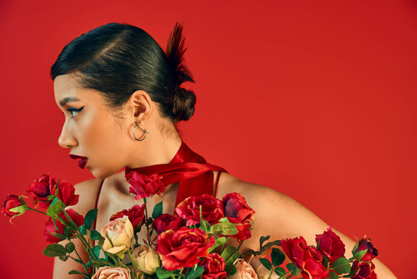 portrait of beautiful asian woman with brunette hair, bold makeup and trendy hairstyle, wearing neckerchief and looking away near bouquet of roses on red background, trendy spring concept - Photo, Image