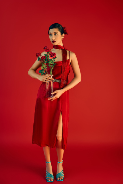 full length of youthful and elegant asian woman in stylish strap dress and neckerchief, with bold makeup and brunette hair standing with roses on red background, trendy spring, generation z - Фото, изображение