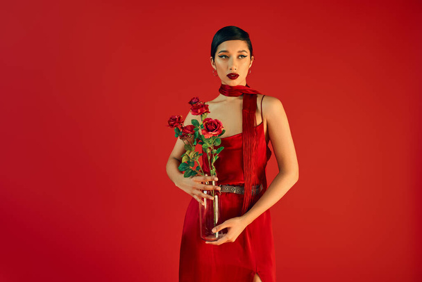 gen z fashion, stylish spring concept, charming asian woman in elegant dress and neckerchief holding glass vase with roses and looking away on red background - Foto, imagen