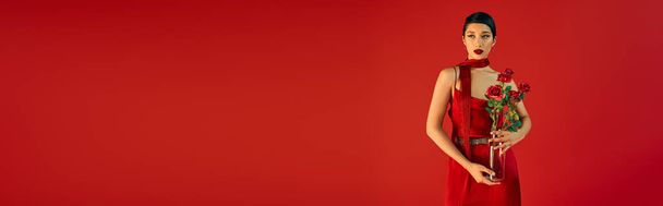 mladá a svůdná asijská žena s tučným make-upem a brunetovými vlasy, v elegantních šatech a krčníku s kyticí růží na červeném pozadí, jarní módní fotografie, banner - Fotografie, Obrázek