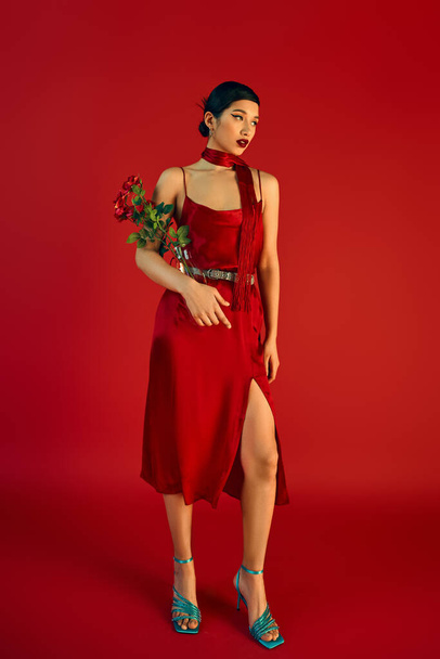longitud completa de atractiva mujer asiática en traje de primavera de pie con flores sobre fondo rojo, vestido de correa elegante, pañuelo, sandalias de color turquesa, sesión de moda - Foto, Imagen
