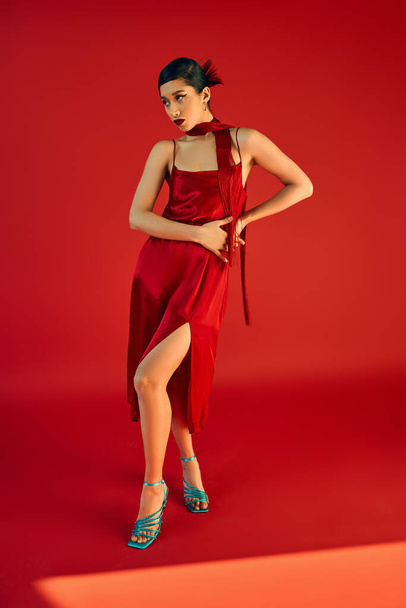 full length of attractive asian woman in elegant dress, neckerchief and turquoise sandals holding hands on hip while standing on red background with lighting, gen z fashion, trendy spring concept - Photo, Image