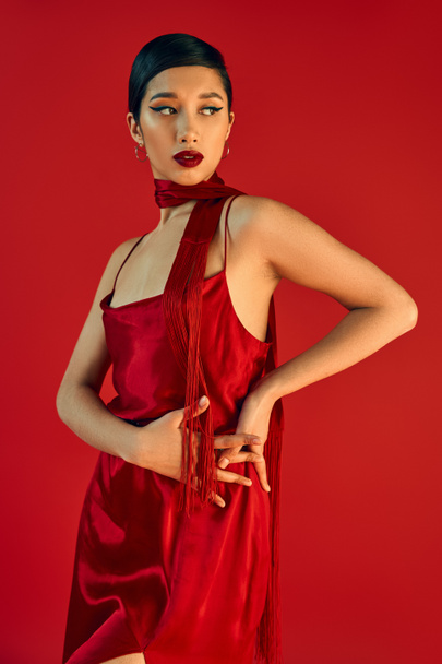 young and fashionable asian woman with bold makeup and brunette hair wearing elegant strap dress and neckerchief while posing with hands on hip on red background, spring fashion concept - Foto, afbeelding