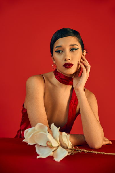 dreamy asian woman with brunette hair and bold makeup holding hand near face and looking away near white orchid on red background, spring fashion, strap dress, stylish neckerchief - Φωτογραφία, εικόνα