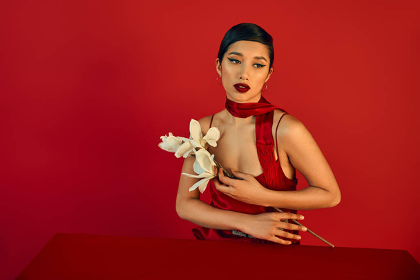 sensual and brunette asian woman with bold makeup, in elegant dress and neckerchief sitting at table with white blooming orchid on red background, generation z, trendy spring concept - Photo, Image