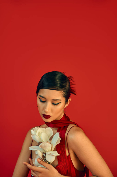 youthful and charming asian fashion model in strap dress and neckerchief, with brunette hair and bold makeup looking at white blooming orchid on red background, trendy spring, fashion shoot - Fotó, kép