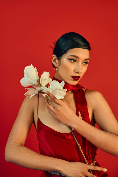 romantic asian woman in spring outfit holding white orchid and looking away on red background, brunette hair, bold makeup, strap dress, neckerchief, generation z - Zdjęcie, obraz