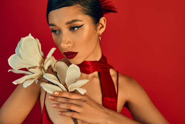 ritratto di giovane e attraente donna asiatica con orchidea bianca in fiore su sfondo rosso, trucco audace, capelli castani, fazzoletto, fotografia di moda primaverile, elegante e affascinante  - Foto, immagini