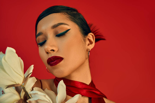 portrait of sensual asian fashion model with brunette hair, bold makeup and trendy hairstyle posing with closed eyes near white blooming orchid on red background, spring fashion, generation z - Foto, Imagem