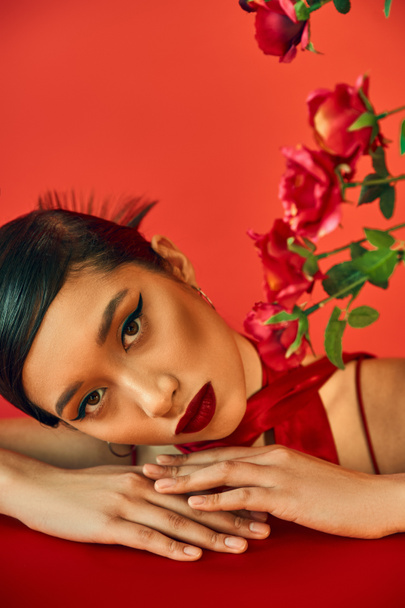 portrait of young and appealing asian fashion model with bold makeup and brunette hair looking at camera while laying on table near roses on red background, stylish spring, fashion shoot - Fotó, kép