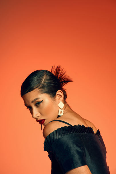 portrait of glamour asian woman with bold makeup and brunette hair, wearing trendy earrings and black elegant dress while posing on orange background, generation z, spring fashion photography - Foto, immagini